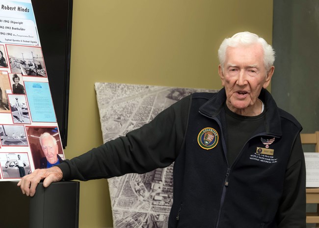 Older man in a volunteer uniform points towards a panel with his historic images.