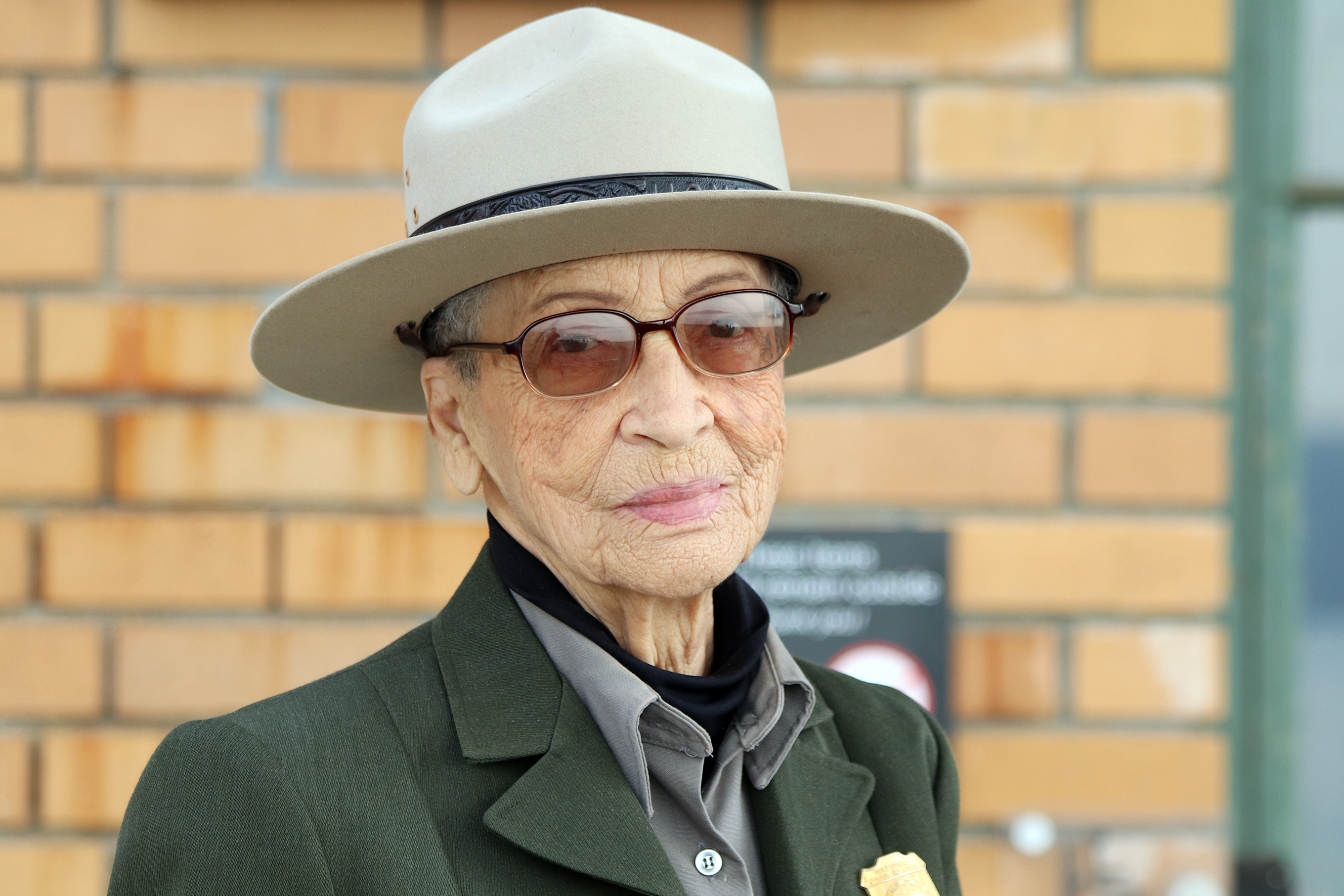 Ranger Betty Reid Soskin pictured in front of the Rosie the Riveter Visitor Center wearing a green and gray National Park Service uniform with sunglasses and a tan flat hat.