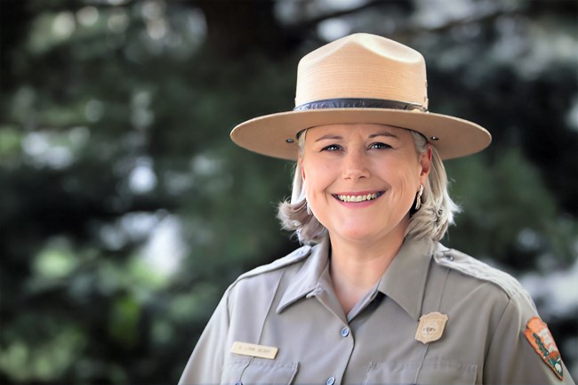 K. Lynn Berry, wearing her National Park Service ranger uniform and hat, smiles warmly. She stands outdoors with a blurred green background of trees.