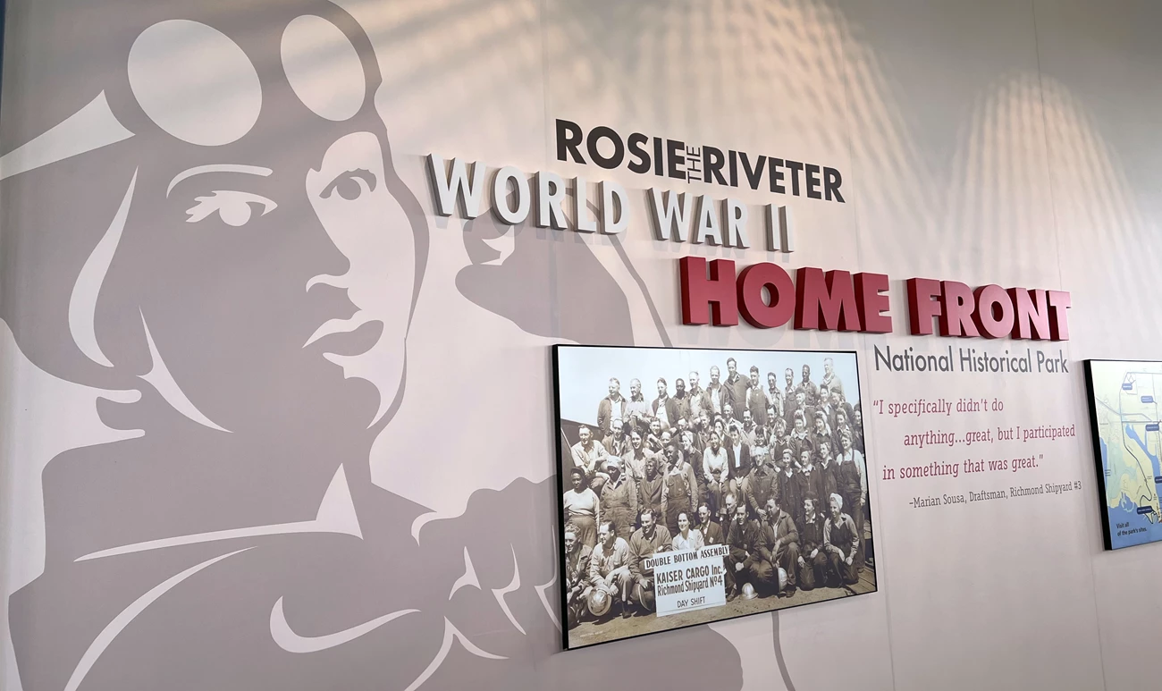 VC entrance wall graphics. Entrance display at the Rosie the Riveter/World War II Home Front National Historical Park showing a large illustration of a female worker representing Rosie the Riveter, a historical group photo of shipyard workers, and a quote from Marian Sousa, a WWII