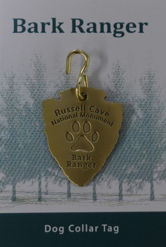 A metal dog tag sits on a paper with trees drawn as a background.  On the top of the paper, the words "Bark Ranger" are written.  On the tag, the text reads "Russell Cave National Monument Bark Ranger"  with a paw print in the center.