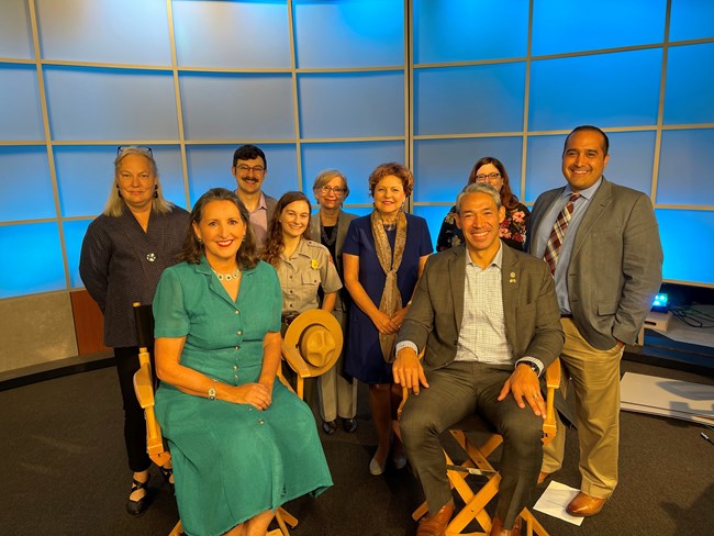 Nine people smile at the camera in a group with blue light backdrop. Included in the picture is the Mayor of San Antonio and a Park Ranger from San Antonio Missions.