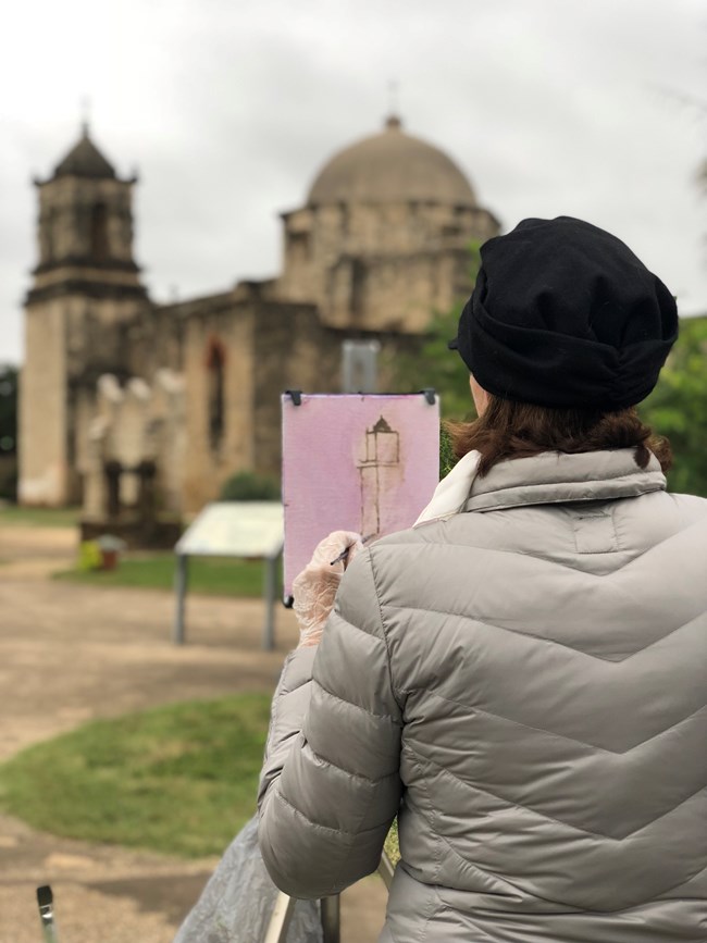 Woman painting Mission San Jose in watercolor while standing in front of her easel at the Mission San Jose church.