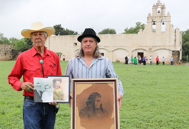 Two men standing in front of Mission San Juan church holding picture frames of their ancestors.