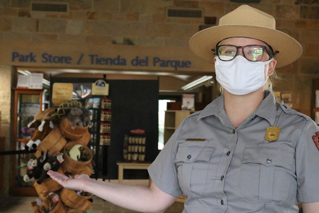 Park Ranger wearing white mask stands in front of stocked Park Store with outstretched arm.