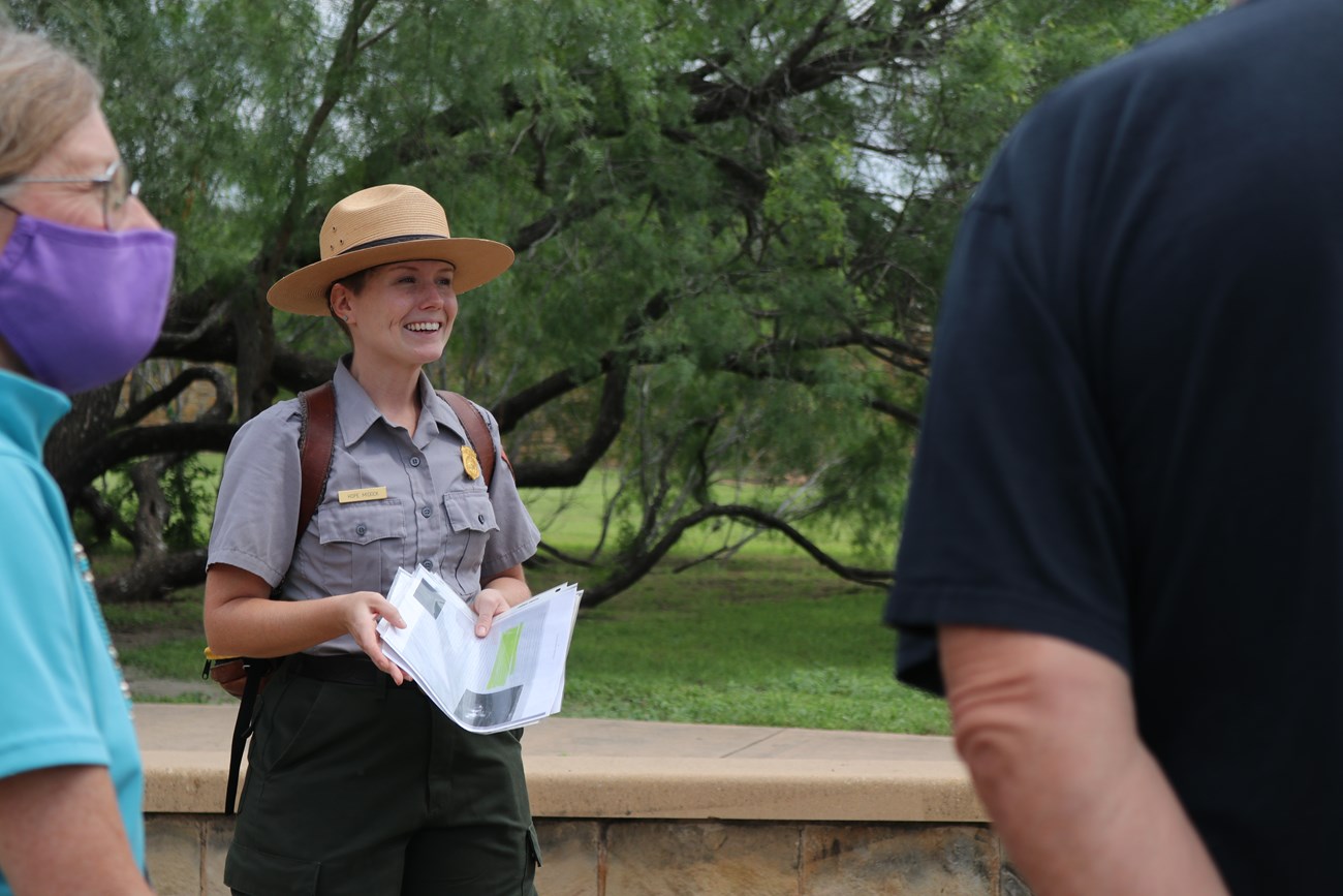 Female park ranger smiles while leading a tour and holding a stack of maps