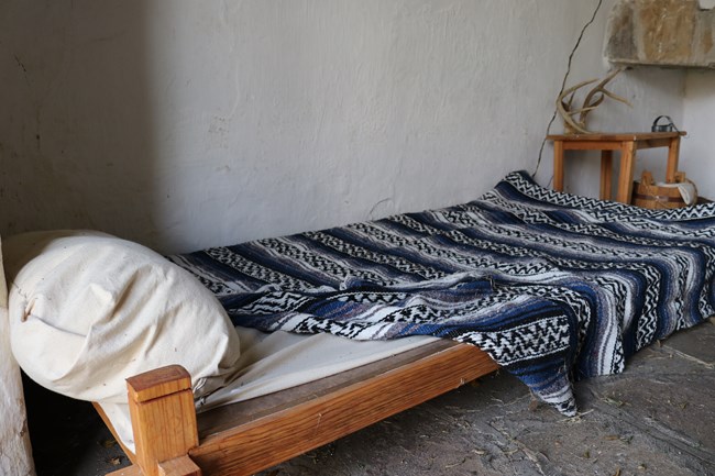 In the foreground, a bed with wooden frame, pillow, and blanket with blue, white, and black design. In the background, a small wooden table with deer antlers on top.