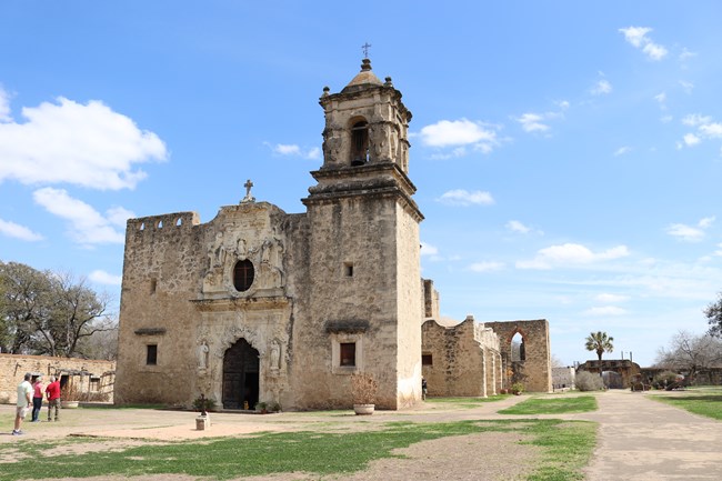 Limestone, intricately carved Mission San Jose church facade with few visitors in foreground on sunny day