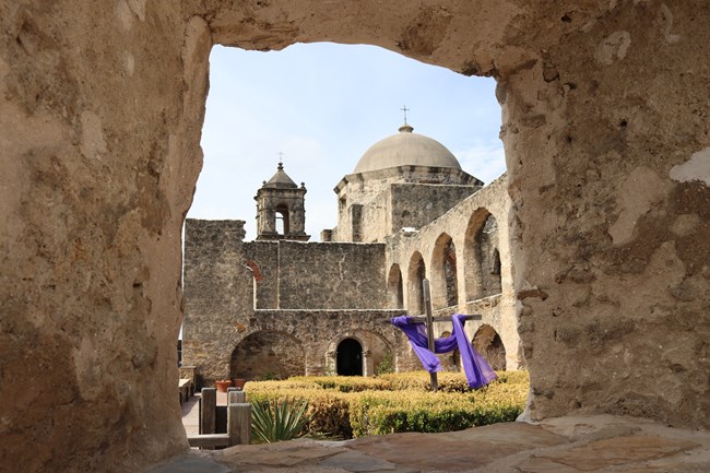 In the foreground, a window in the convento.  In the background, a view of the convento stone arches, and Mission San Jose stone church.