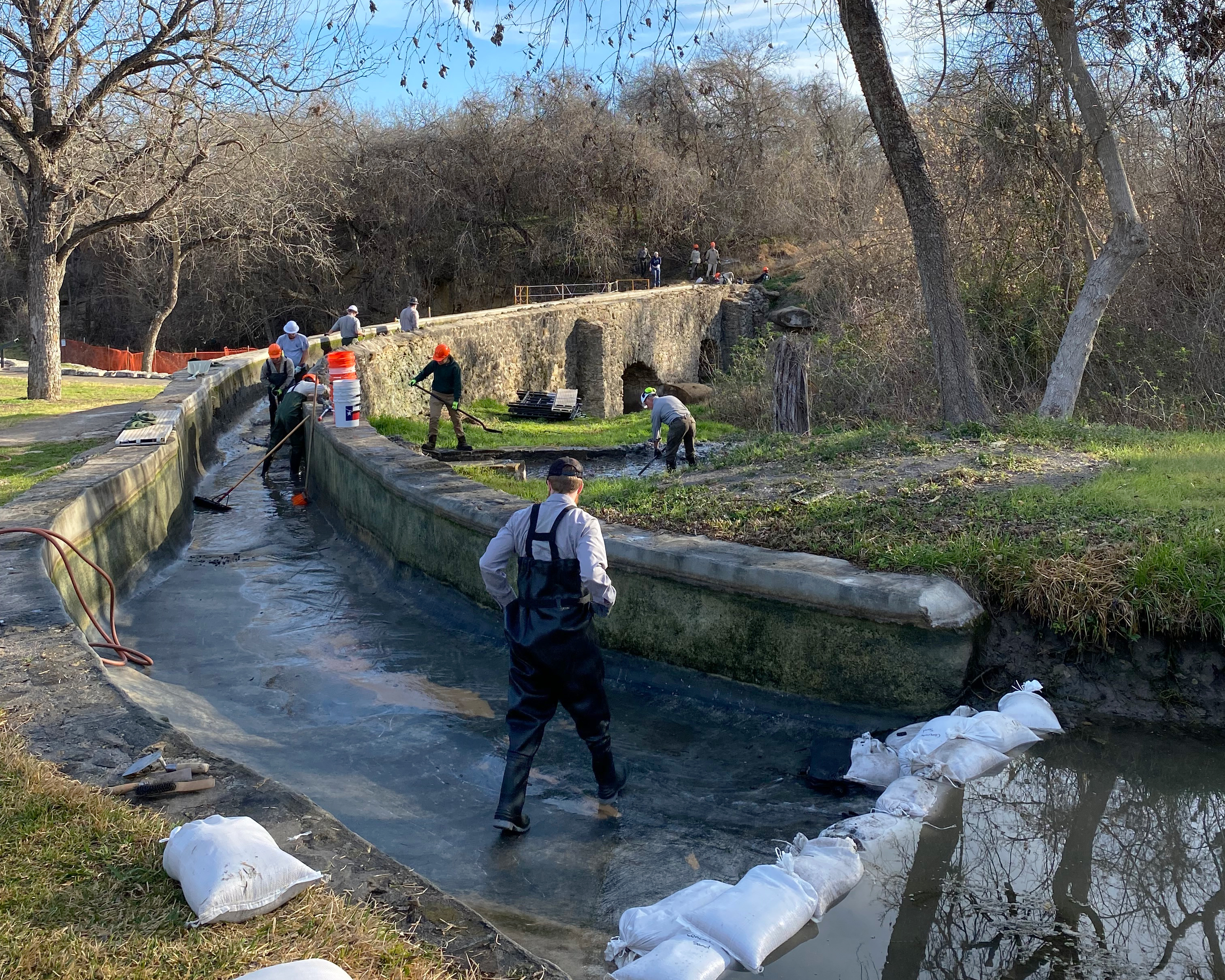 Espada Aqueduct Repair HPTC workers in hard hats stand in a narrow channel of a stone structure with brooms