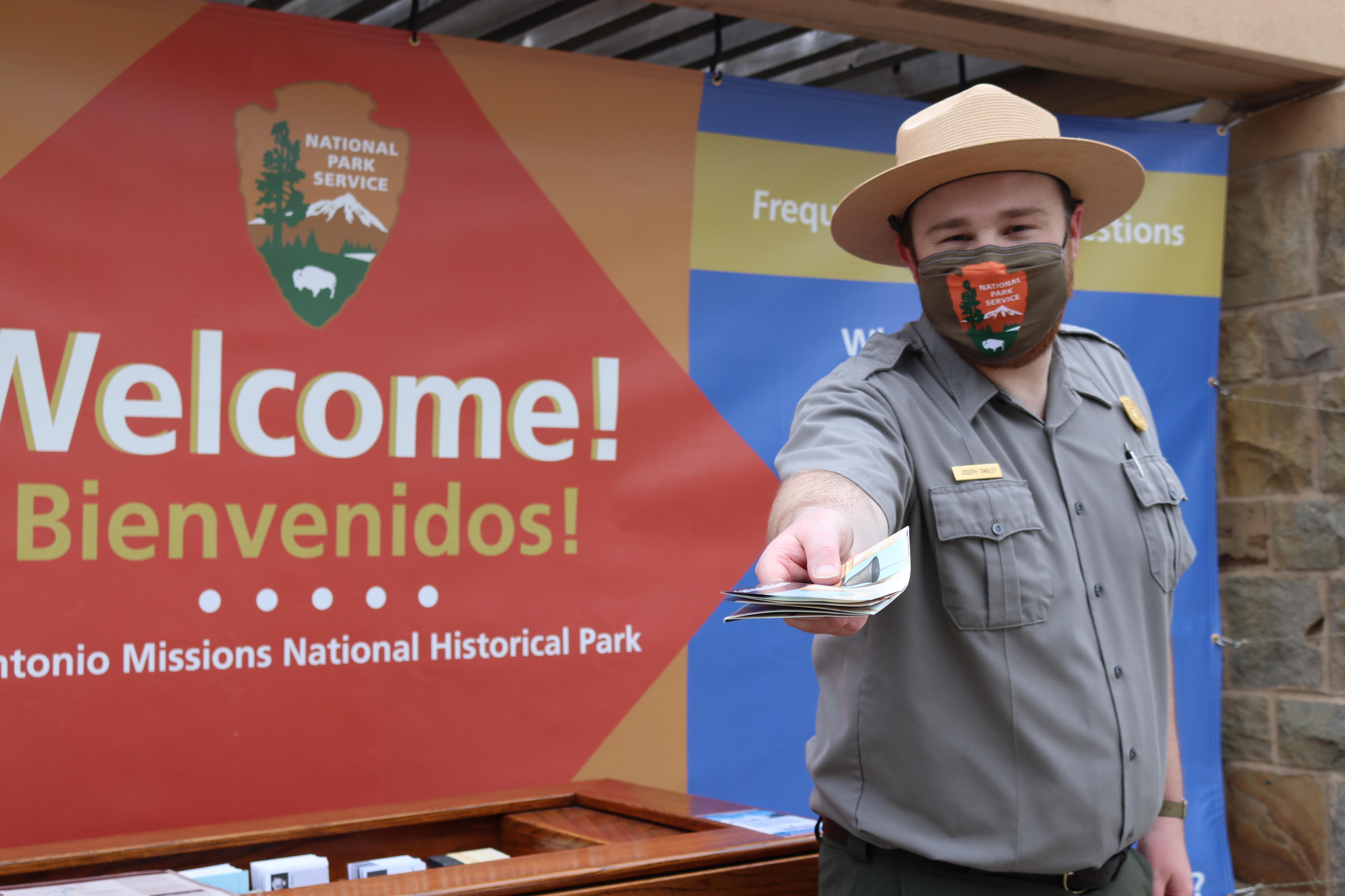 Masked Park Ranger holds park brochure in front of large welcome banner.