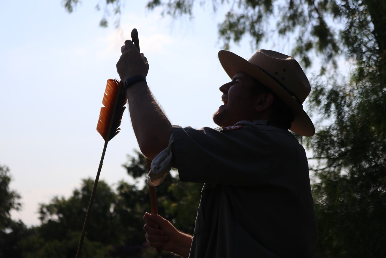 Silhouette of Park Ranger holding an atlatl and dart. Sunlight reflects off of the red feathers at the top of the dart. Outdoors.