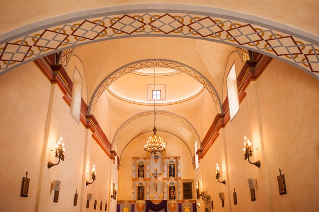 Interior of mission church, looking down pews with light fixtures above.