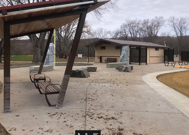 Concrete plaza waiting area with wood and metal shelter, benches (two wooden, two concrete, and three are rocks), and two tall exhibits (wrapped in plastic).  Cream and brown colored comfort station in the background with brown shield corners at doors.
