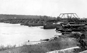 A log dam stretches across the river with many piers and a center gate.  A road runs across the top