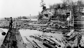 With rocky bluffs in background floating logs and men standing on logs in foreground