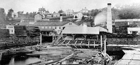 logs, cut boards and bulidings with large chimneys are in front with houses on the hill in back
