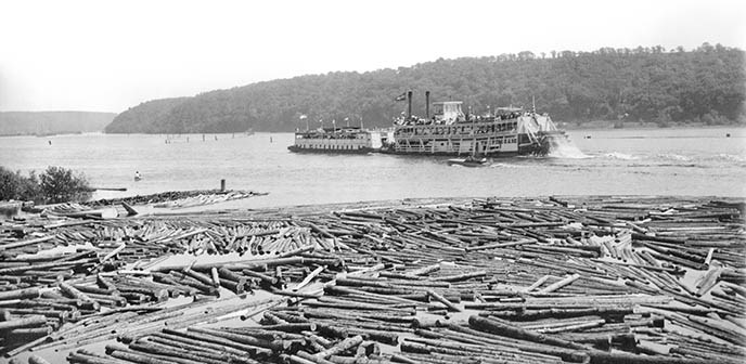 A large paddle-wheel and smaller boats motor on a river filled with logs in the foreground.