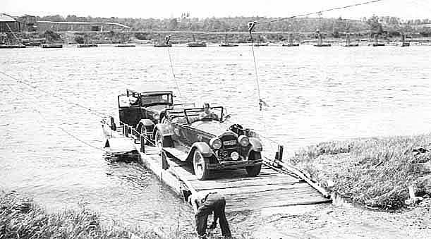 Wooden railroad bridge is in background.  Two cars on a wooden platform in the river are in the foreground