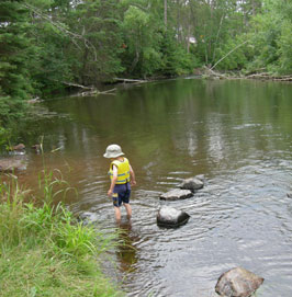 A little boy with a white hat and a yellow lifejacket is wading in the tree lined and rocky river A little boy with a white hat and a yellow lifejacket is wading in the tree lined and rocky river