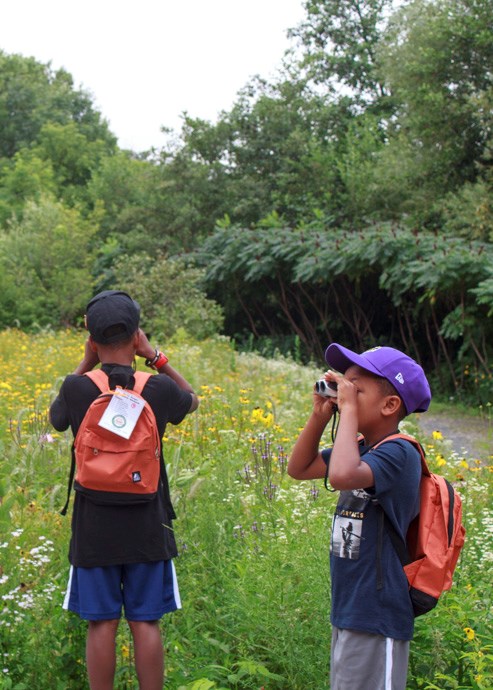Two boys wearing backpacks look through binoculars in a garden