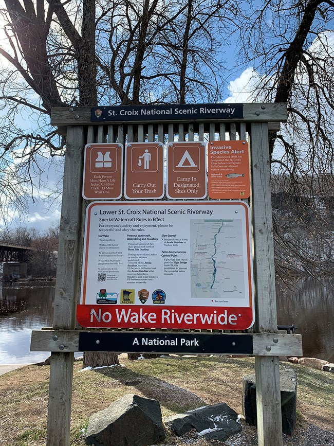 Wooden bulletin board with metal white on brown signs for wearing life jackets, carrying out trash, camping in designated sites, and watching for invasive species.  Below these is a large metal sign with a map of the river and list of boating regulations.