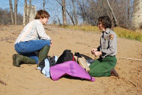 A man in jeans and a shirt and a woman in the gray and green national park service field uniform kneel and talk on a sandy shore.