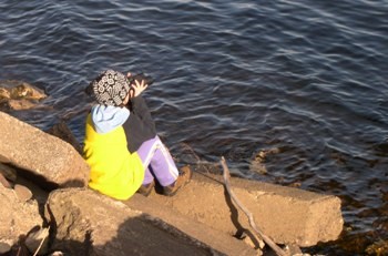 A person in a yellow jacket looking through binoculars, sitting on rocks next to the water.