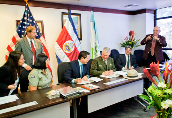 U.S. and Costa Rican flags are displayed behind desks at which two men sign documents, while others look on.