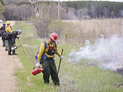 Firefighter with a red canister stands beside a road and other firefighters.