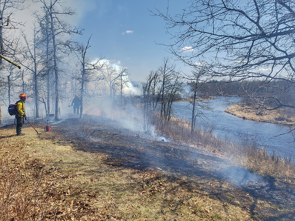 Strip of burning ground along river with rising smoke and two firefighters with equipment
