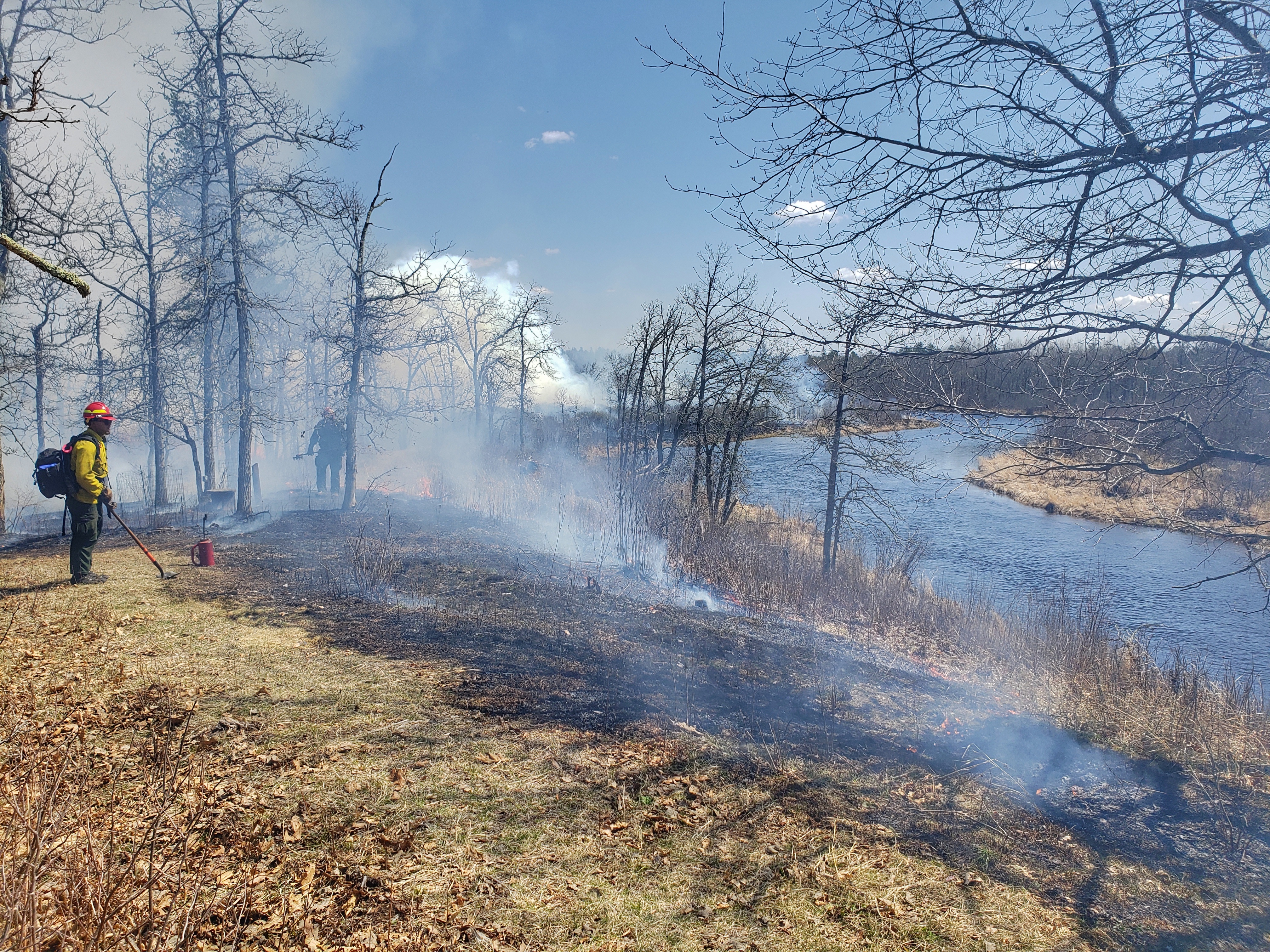 Two firefighters stand near smoking, charred ground next to the St. Croix River.