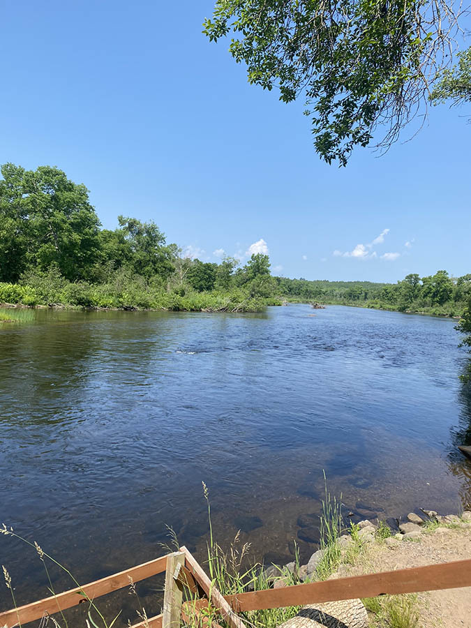 Blue river with green trees in background, blue sky above, and wooden handrail in forefront