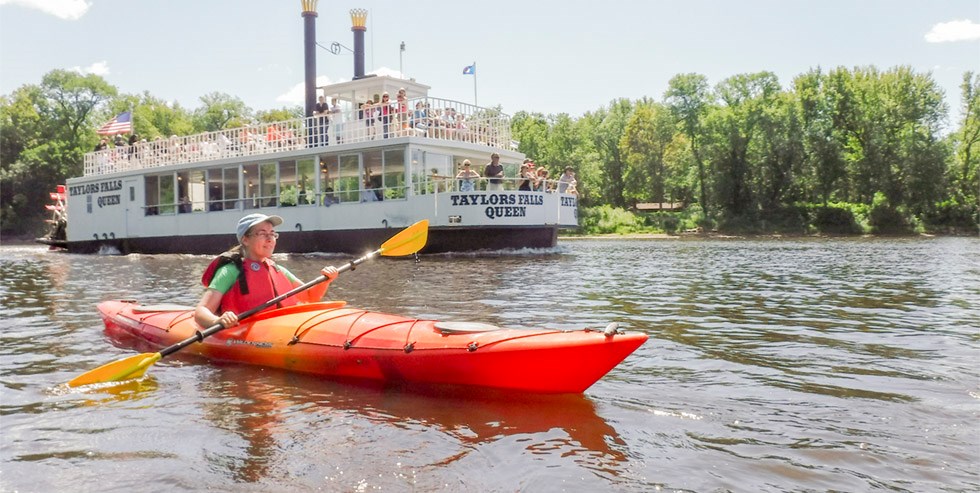 A kayaker paddles near a paddleboat on a river.