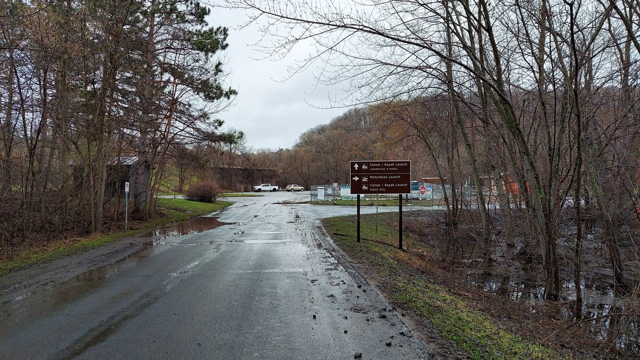 Image of a paved road leading to a parking lot and river access. Road signs and posts line the road's edges.