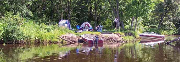 Tents are set up next to a river with boats along the shoreline. Tents are set up next to a river with boats along the shoreline.