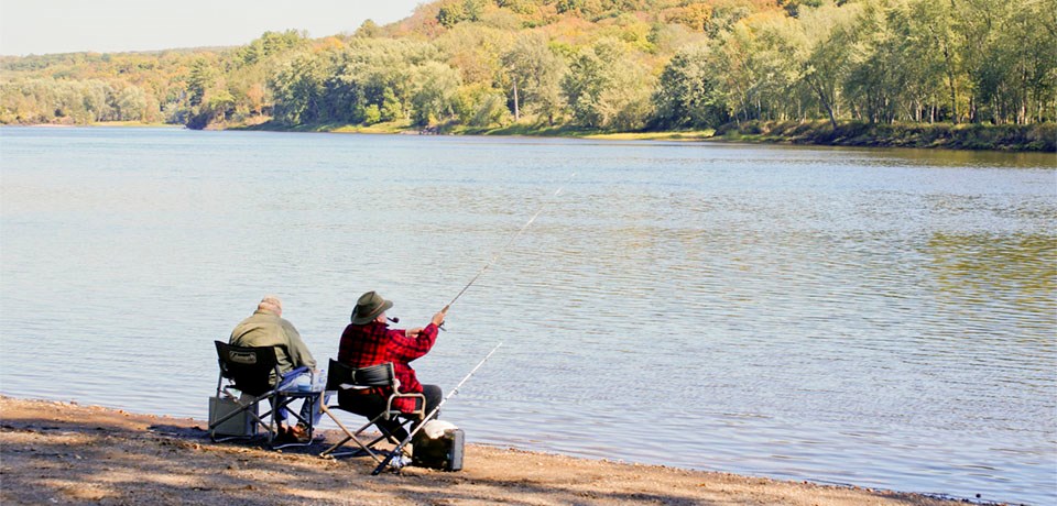 Two men sit in chairs fishing at a river.