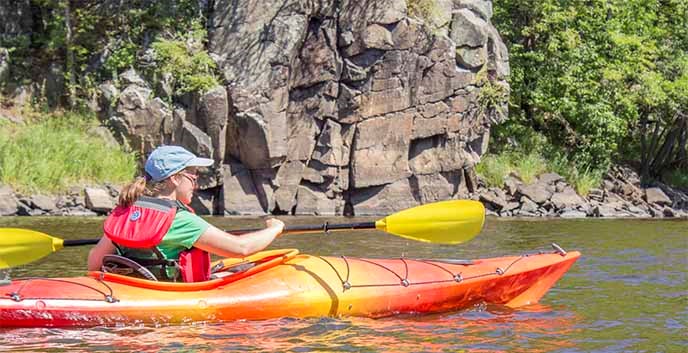 A woman paddles a bright kayak next to a cliff.