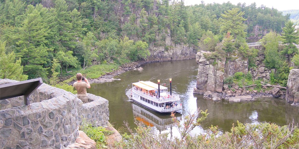 A man takes a picture of a paddleboat on a river.