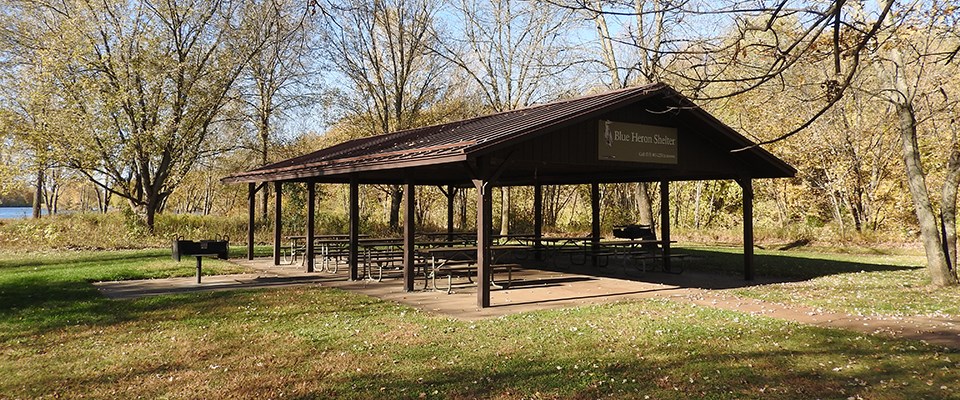An open-sided picnic shelter with picnic tables on a concrete slab sits under a canopy of maple trees in autumn.