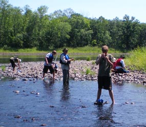 People wading in a shallow river by a rocky river bank, with jars and quizzical expressions