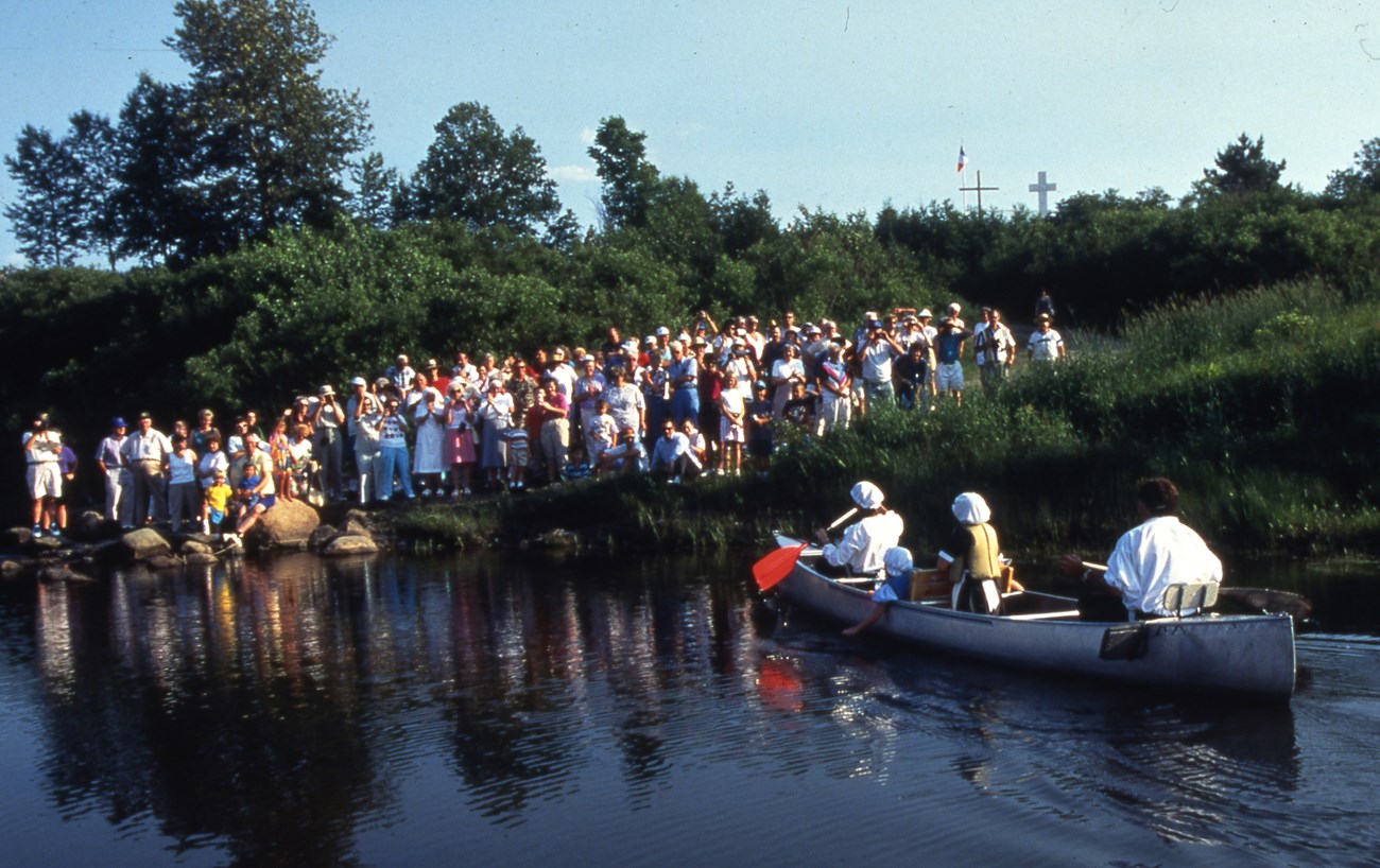 A crowd of people waits on a shoreline while a canoe paddled by people in old-fashioned clothing approaches.