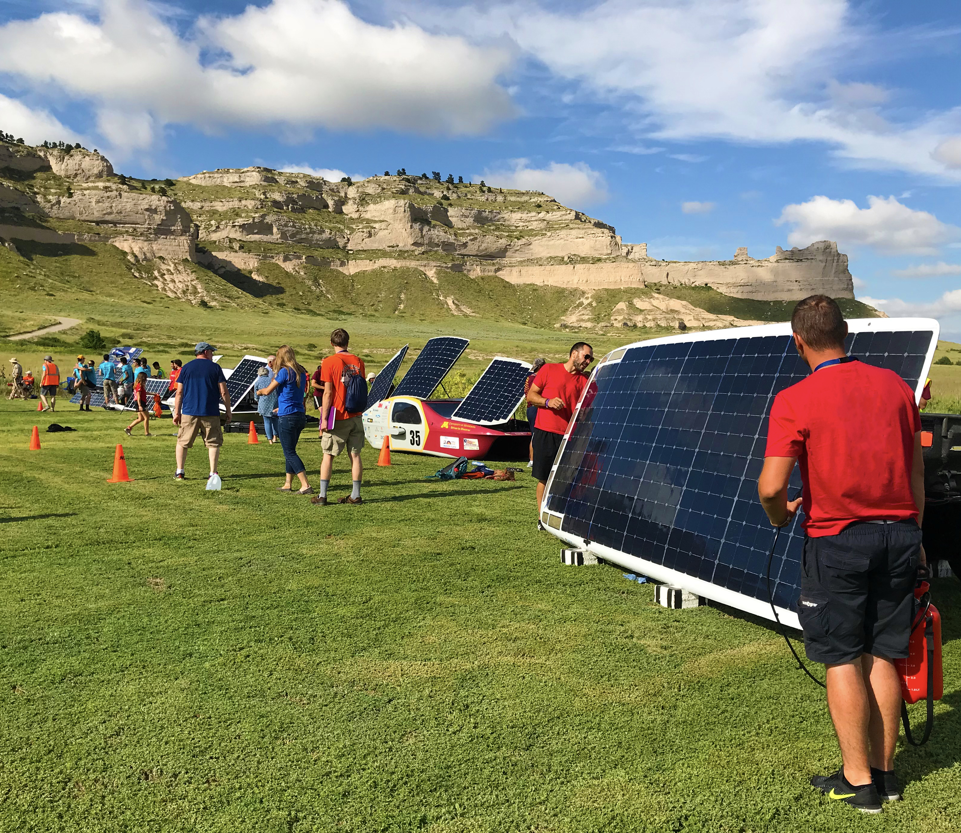 Solar cars propped up for charging on short grass with people milling about and working on the cars. A large bluff and blue skies dotted with clouds are in the background.