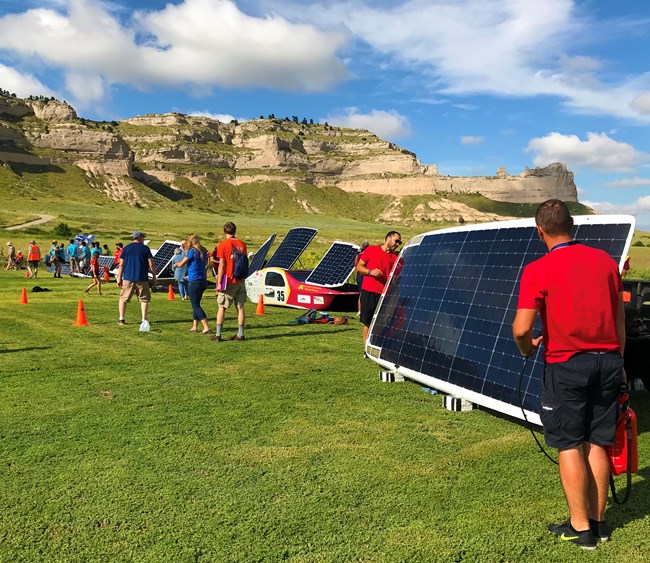 Solar cars propped up for charging on short grass with people milling about and working on the cars. A large bluff and blue skies dotted with clouds are in the background.