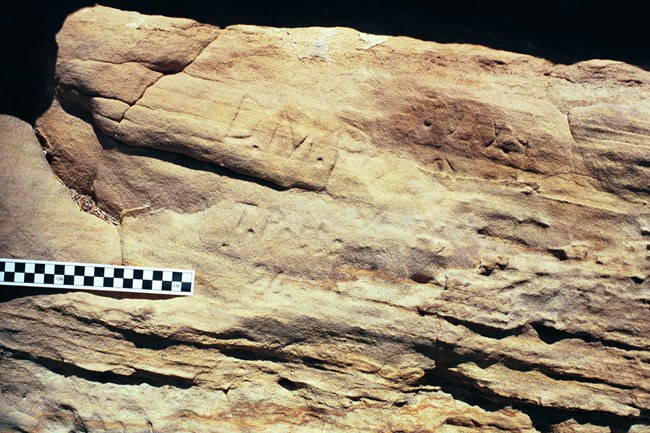 lt= Rough orange and brown-motled slab of sandstone with three faint inscribed names including T.Potts 26. Black and white checkered photo scale on left