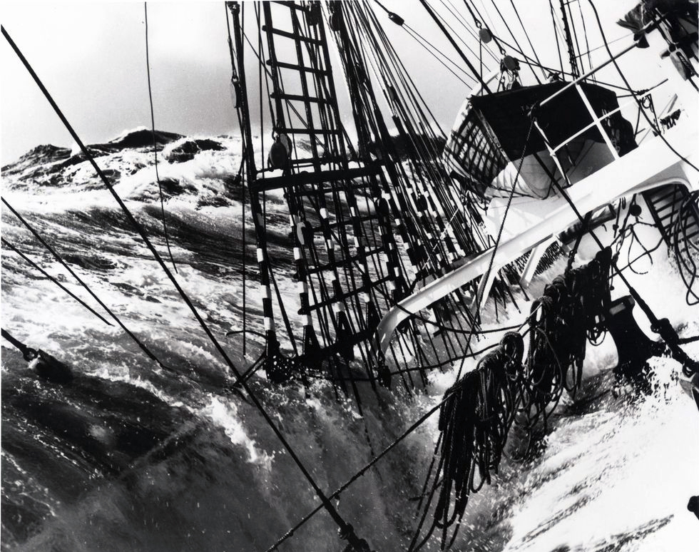 A sailing ship, tilting steeply as it sails in heavy weather. A wave breaks over the rail. The ocean towers above the ship and the water cascades across the deck.