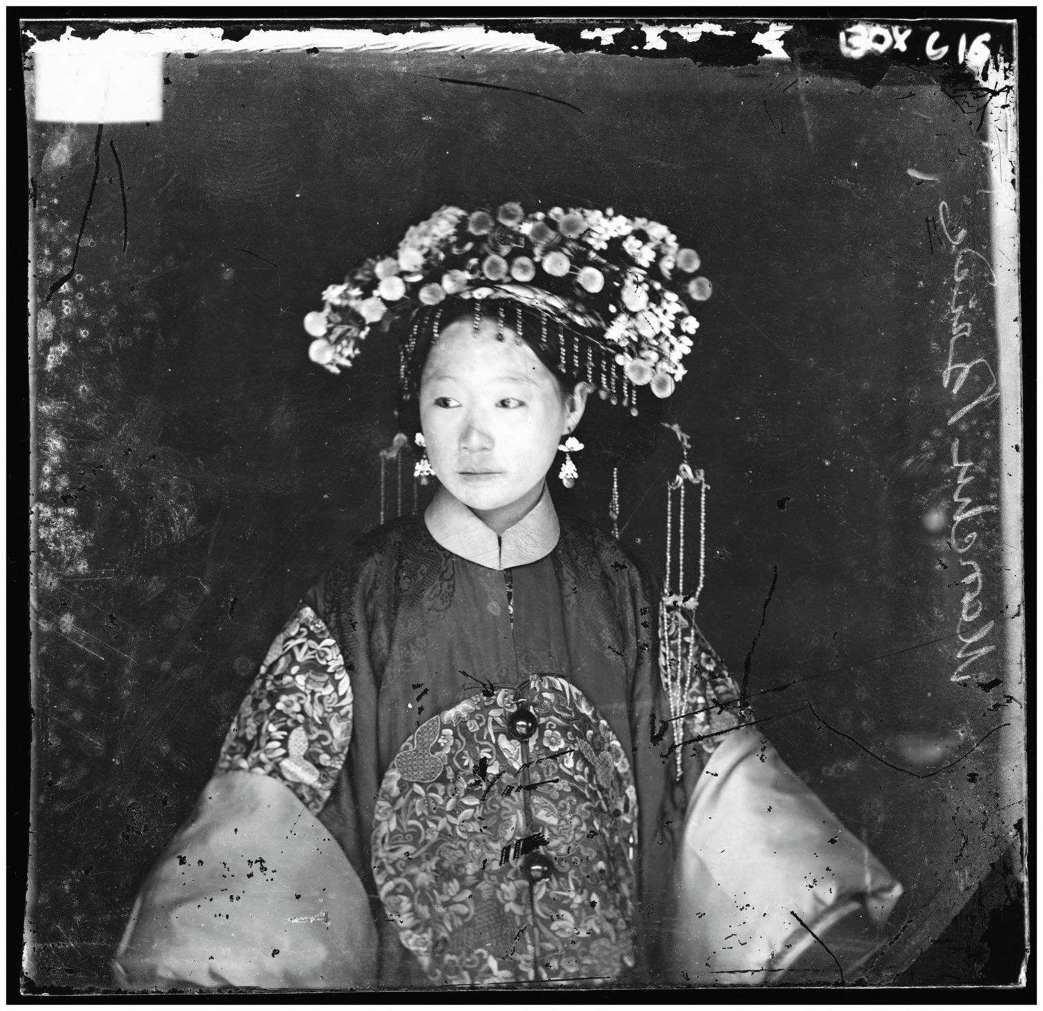 Black and white photograph of a young Chinese bride in elaborate wedding dress. Her face is powdered white, and she is dressed in an intricately embroidered, multicolored silk robe. She wears dangling 
