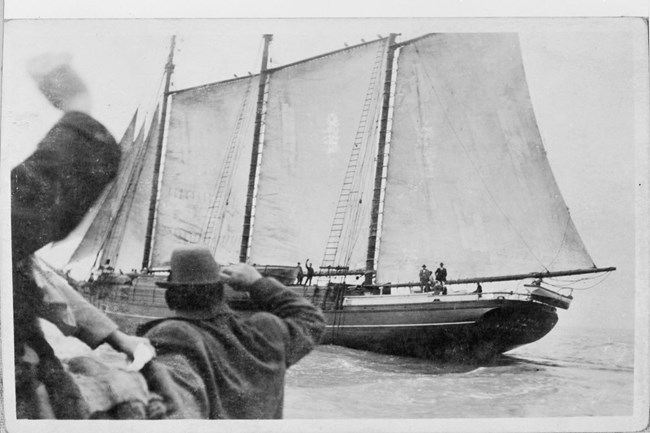 Historical photo of C. A. Thayer setting sail while men cheer from the shore in the foreground.