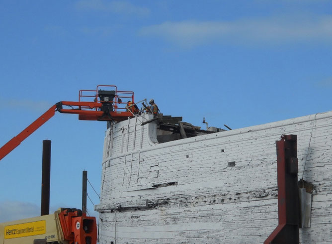 An orange crane positioned over the bow of a wooden-hulled vessel.