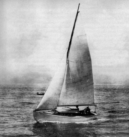 A black and white photo of a wooden sailboat with the mainsail and jib raised.